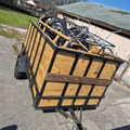 Tilted utility trailer loaded with scrap metal and wood panels on a driveway beside a house.