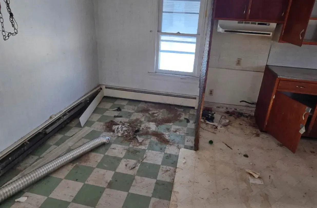 Damaged kitchen with torn floor tiles, debris, and exposed pipes near a window and cabinets