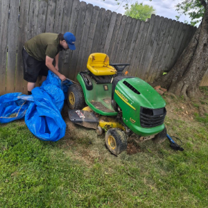 Person covering a green lawn tractor with a blue tarp beside a wooden fence in a backyard