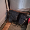 Broken black electronic device and cables lying on a wooden floor in a corner by a wall and door.