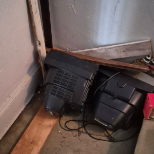 Broken black electronic device and cables lying on a wooden floor in a corner by a wall and door.