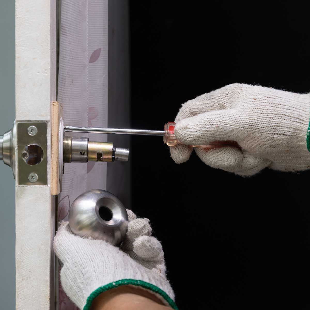 A person wearing gloves is fixing a door knob with a screwdriver