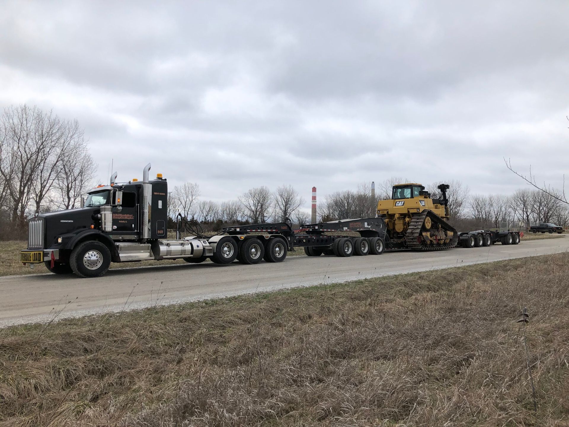 Heavy black semi truck hauling construction equipment on a roadside under a cloudy sky