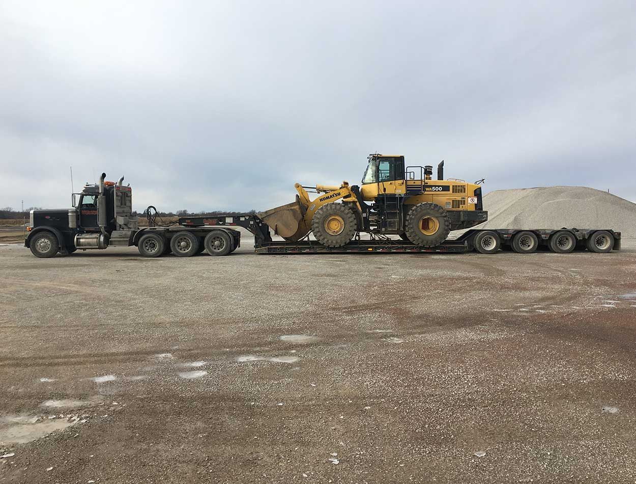 Heavy-duty truck hauling a large yellow loader on a flatbed trailer in a gravel lot under cloudy skies