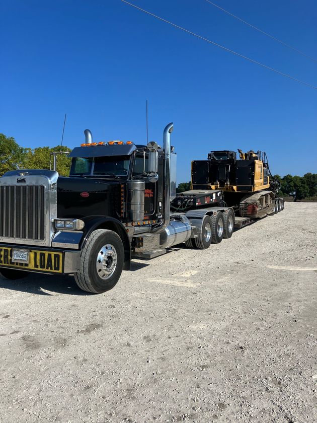 Black semi truck hauling heavy equipment on a flatbed trailer along a gravel road under a clear blue sky