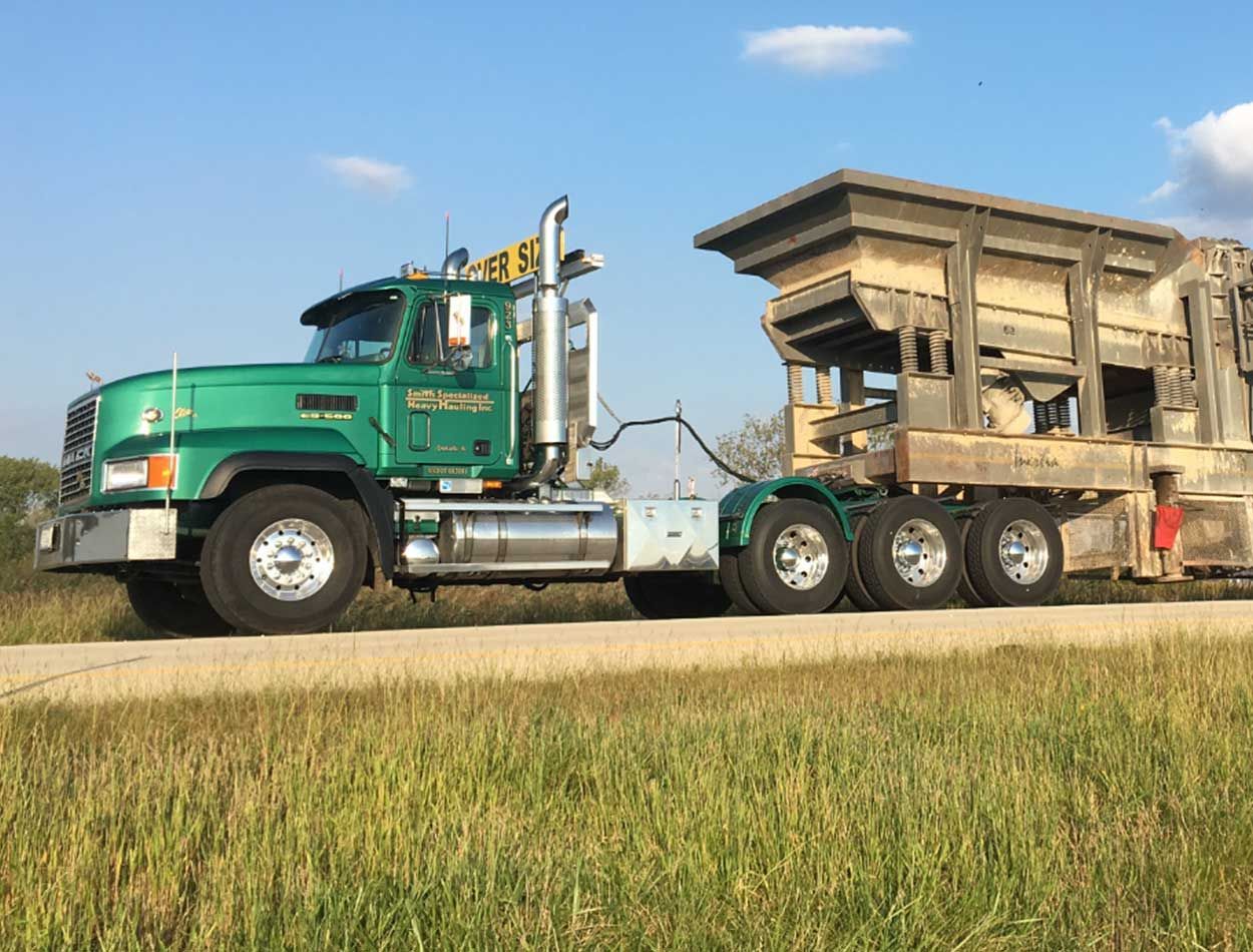 Green semi-truck hauling a large industrial machine along a rural road under a blue sky.