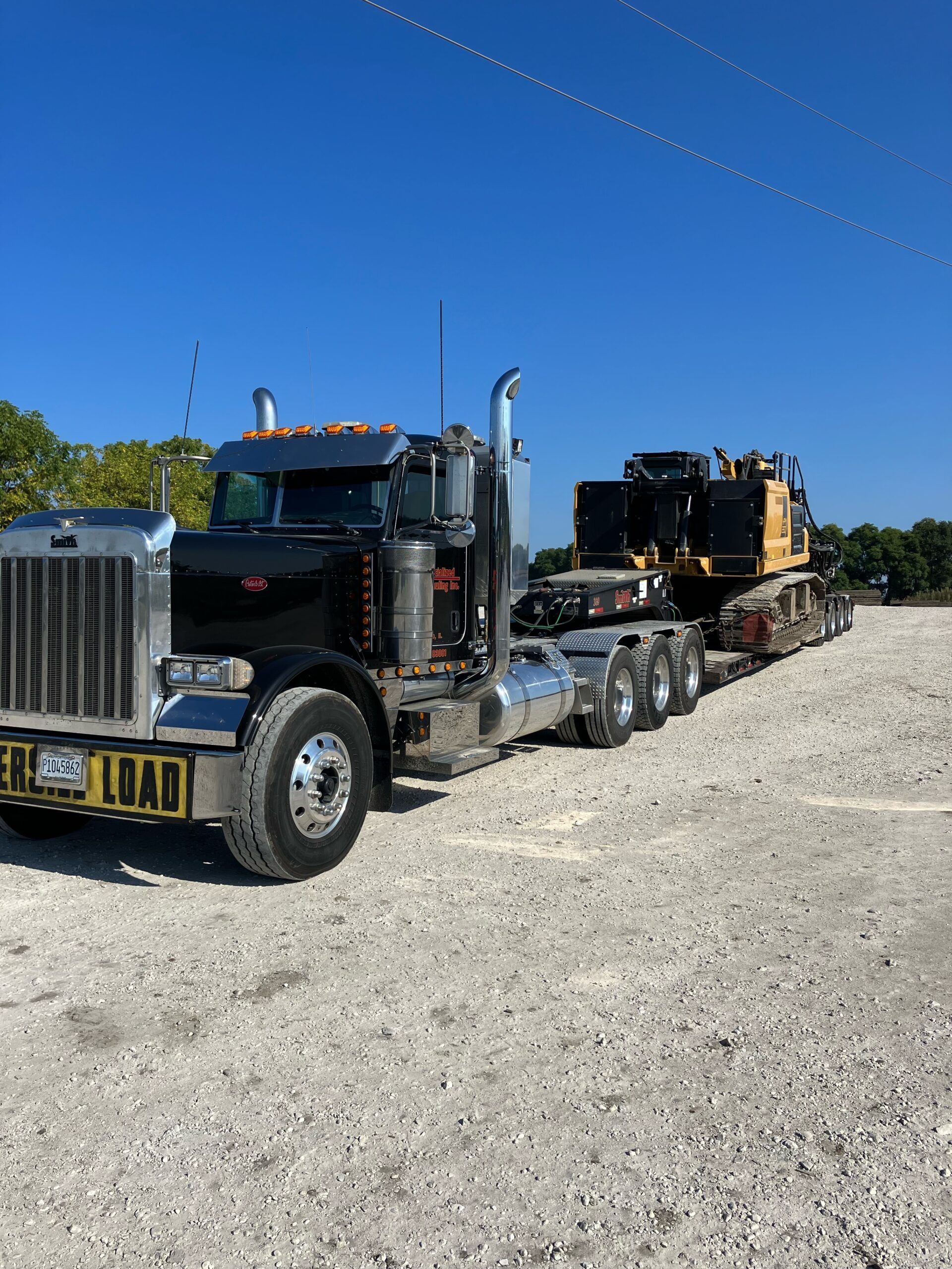 Black semi-truck hauling heavy machinery on a flatbed trailer under a clear blue sky.