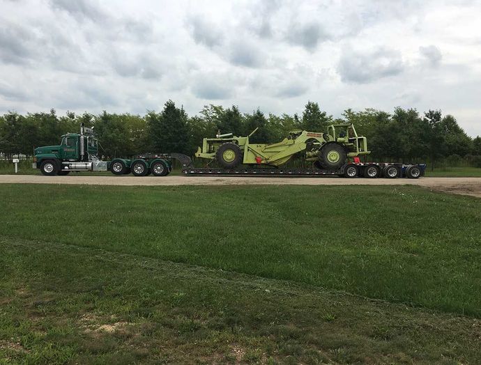Green tractor hauling a yellow farm machine on a flatbed trailer beside a grassy field