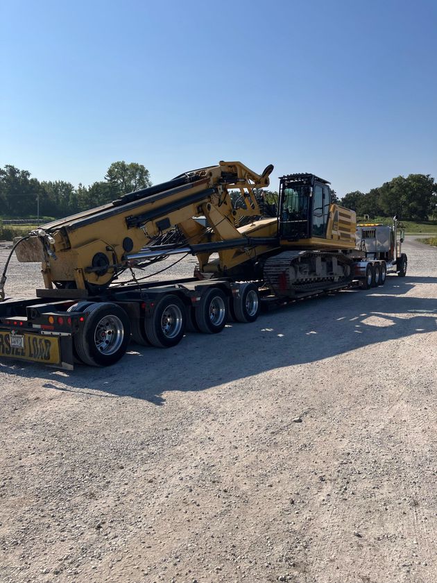 Yellow construction machine loaded on a flatbed trailer in a gravel lot under a clear blue sky.