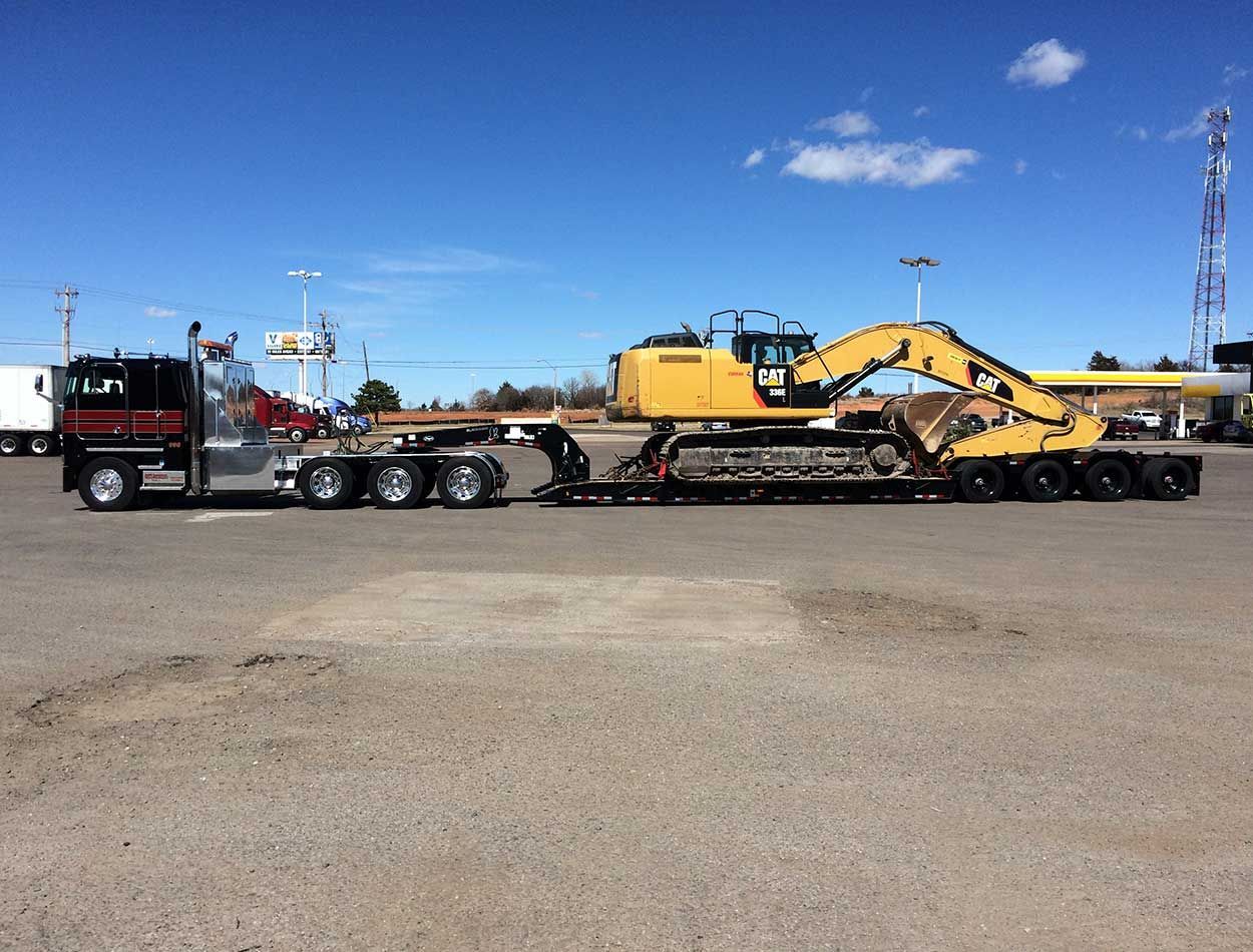 Truck hauling yellow excavator on a flatbed trailer in an open gravel lot under a blue sky