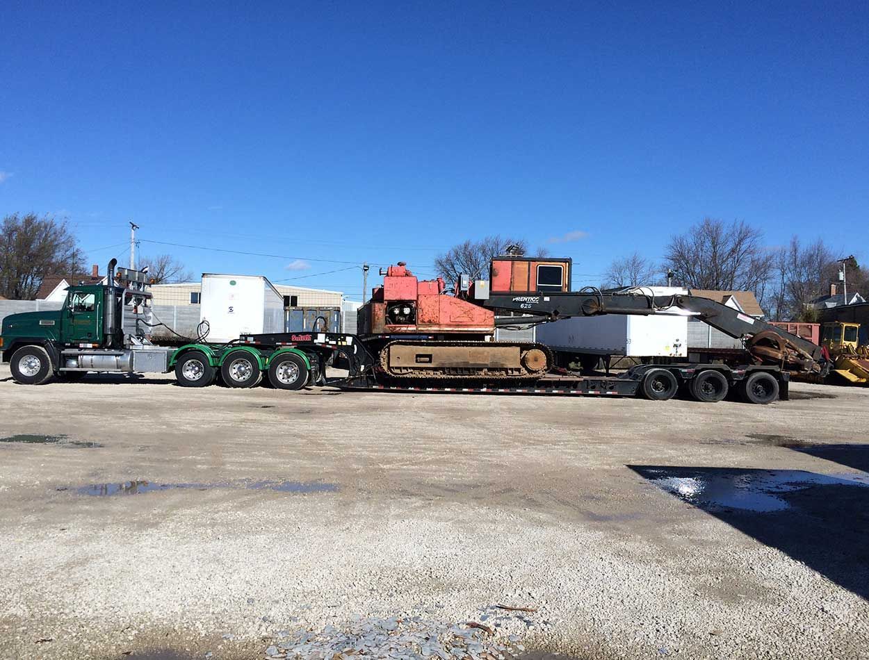Long flatbed trailer hauling heavy equipment in a gravel lot under a clear blue sky
