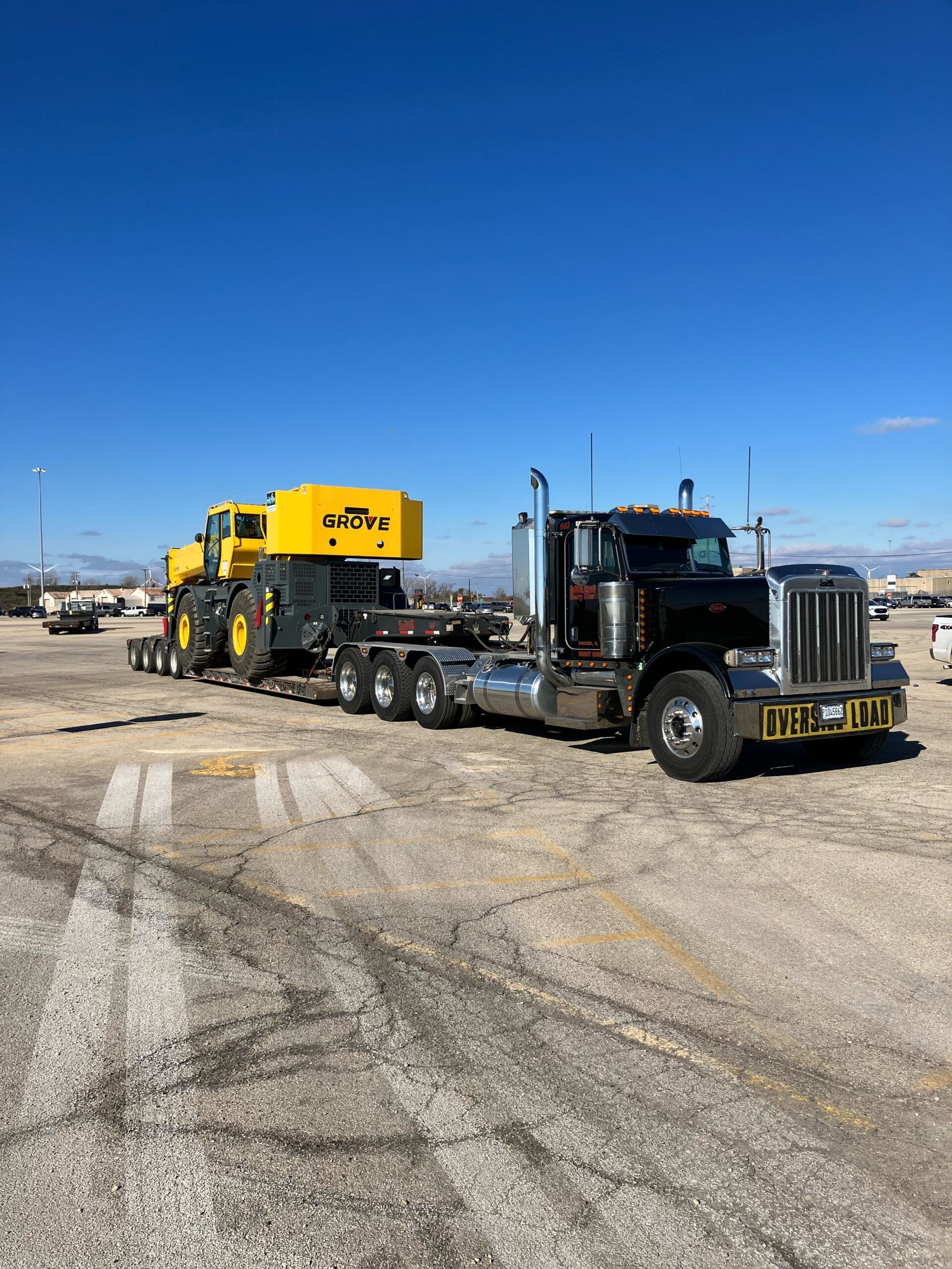 Black semi truck hauling yellow construction machinery on a flatbed in a dusty lot under a blue sky