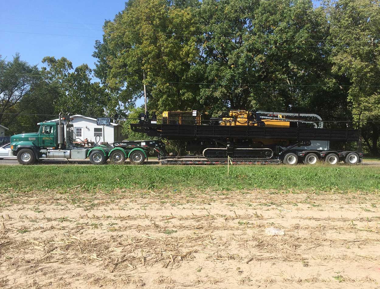 Green semi hauling a long flatbed trailer with heavy machinery along a rural field edge.