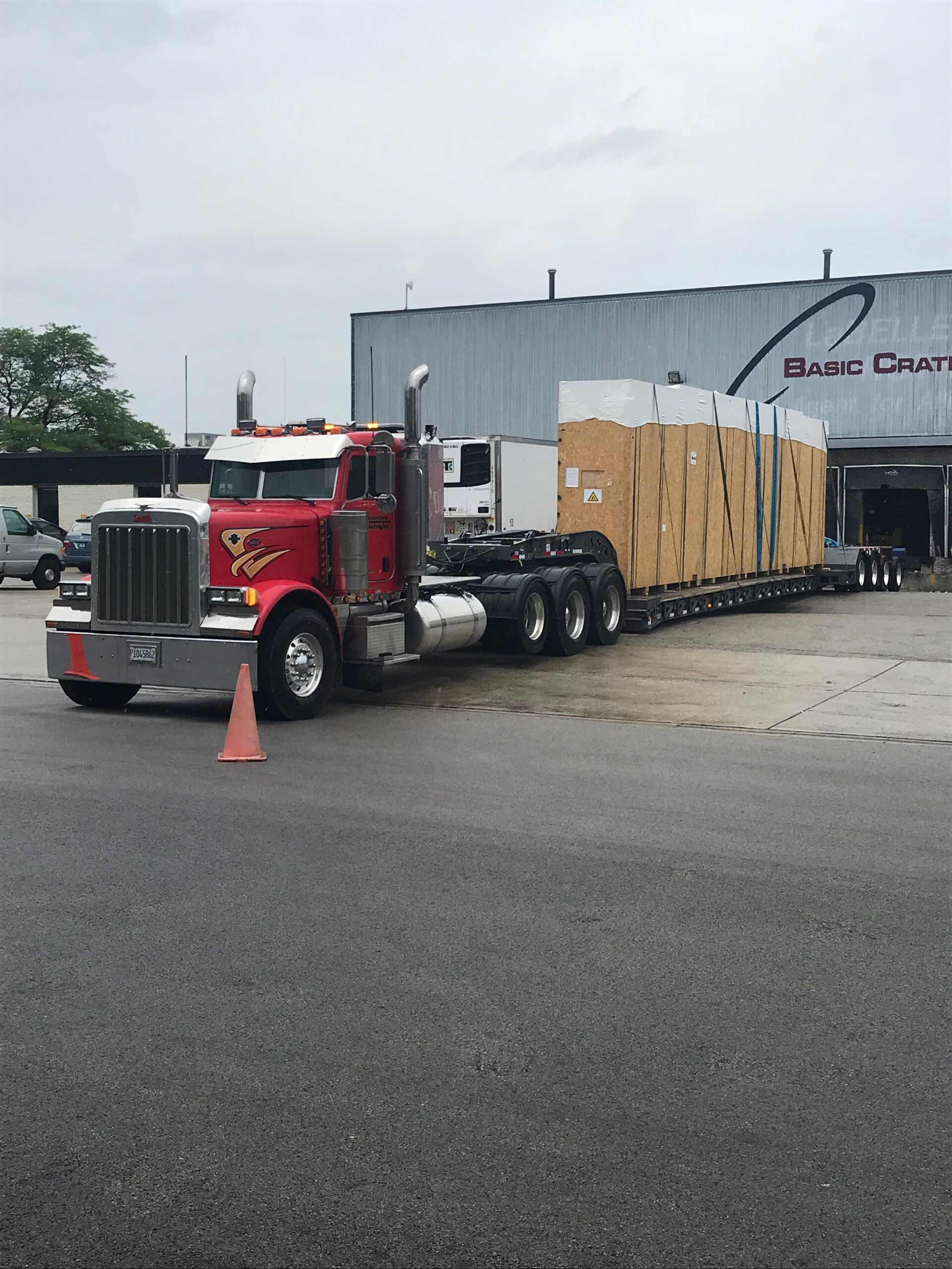 Red semi truck hauling a large trailer parked beside a warehouse on a wet lot