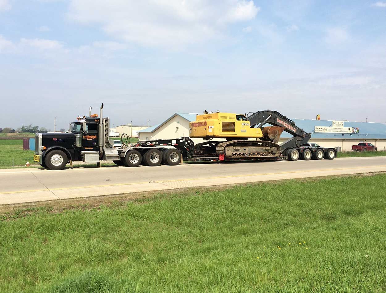 Black semi-truck hauling yellow heavy equipment trailer on a road beside grass and water