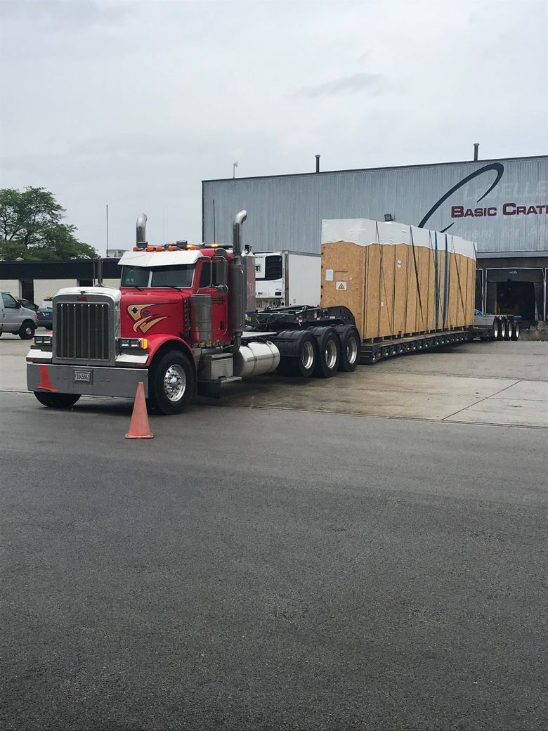Red semi-truck hauling a large wooden crate on a flatbed trailer in an industrial lot.