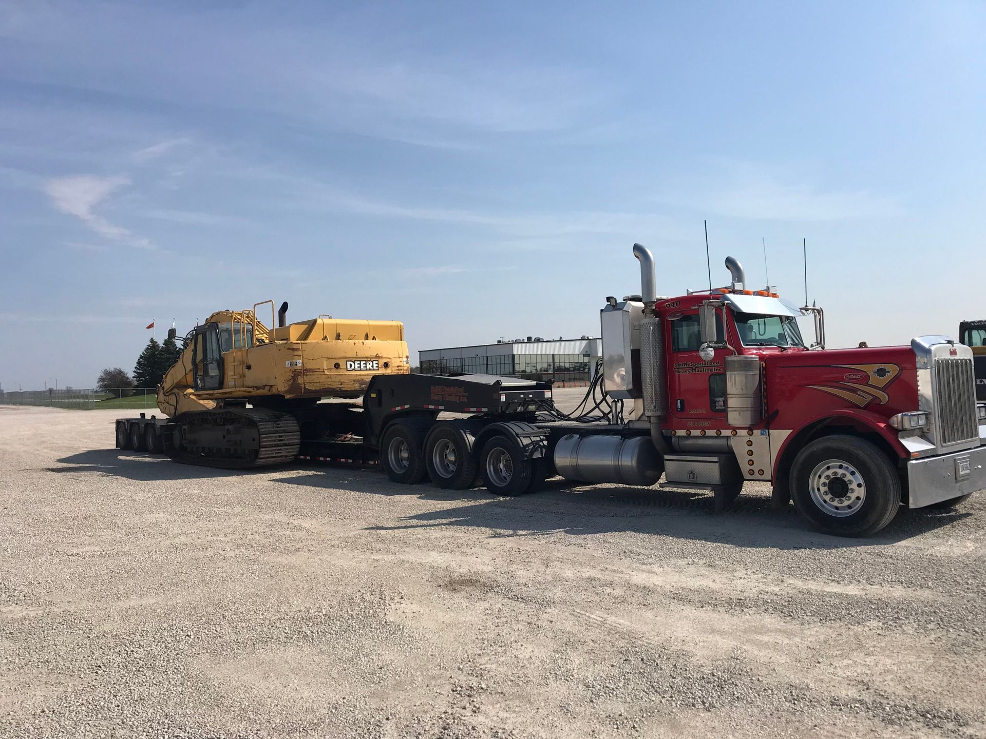 Red semi truck hauling a yellow excavator on a lowboy trailer in a gravel lot.