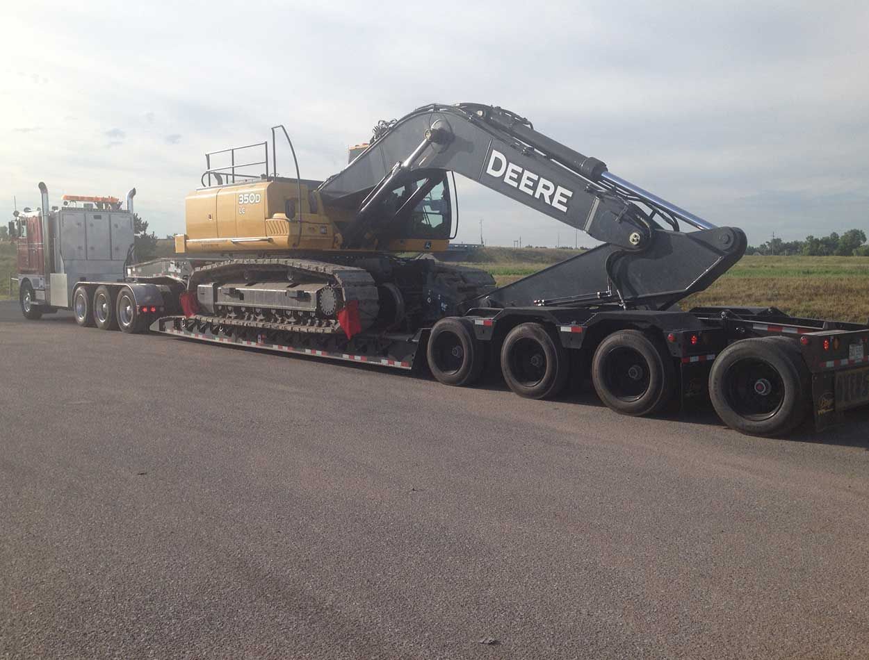Heavy John Deere excavator on a lowboy trailer being transported on a road.