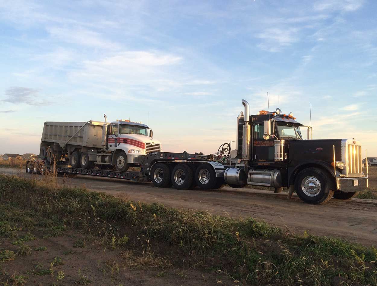 Large semi-truck hauling a dump trailer on a dirt road at sunset, with another truck in the background