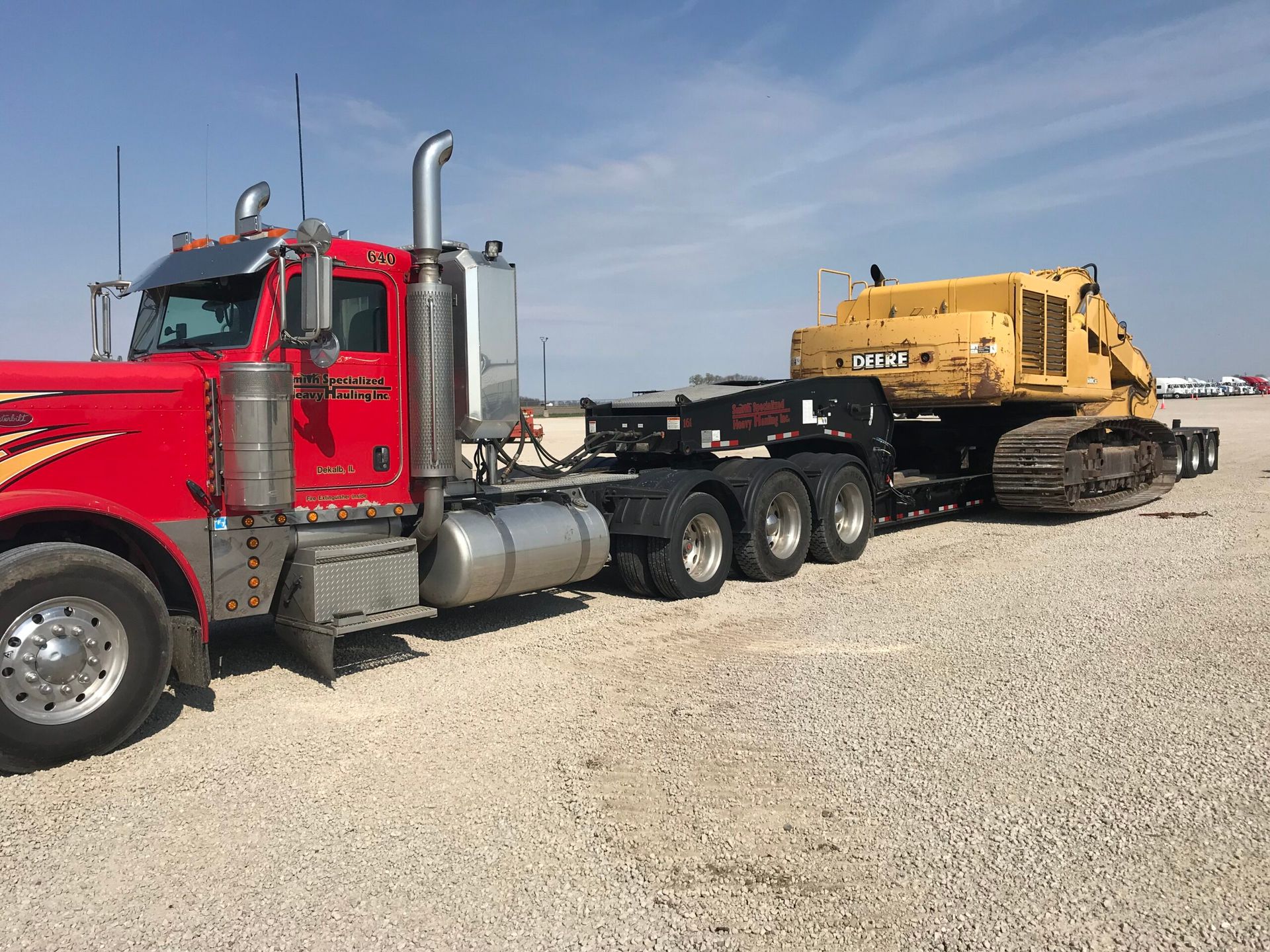 Red semi truck hauling a large yellow construction machine on a flatbed trailer in a gravel lot.