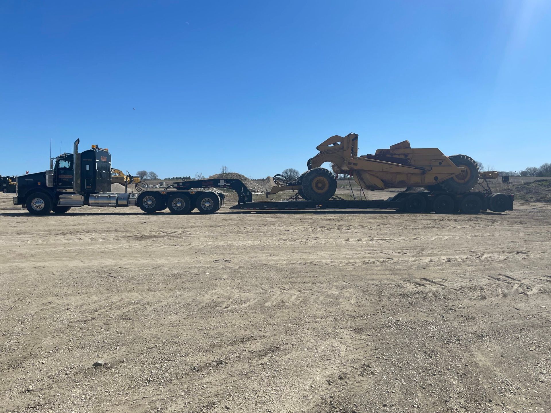 Flatbed truck hauling a large tan bulldozer across a dusty lot under a clear blue sky