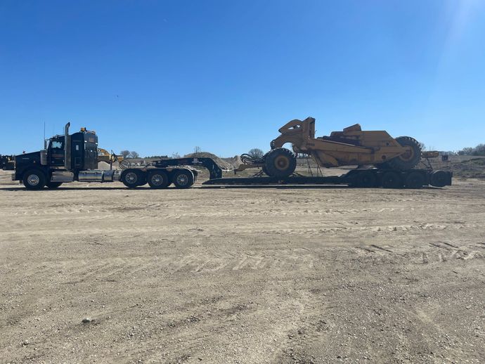 Flatbed truck hauling a large tan bulldozer across a dusty lot under a clear blue sky