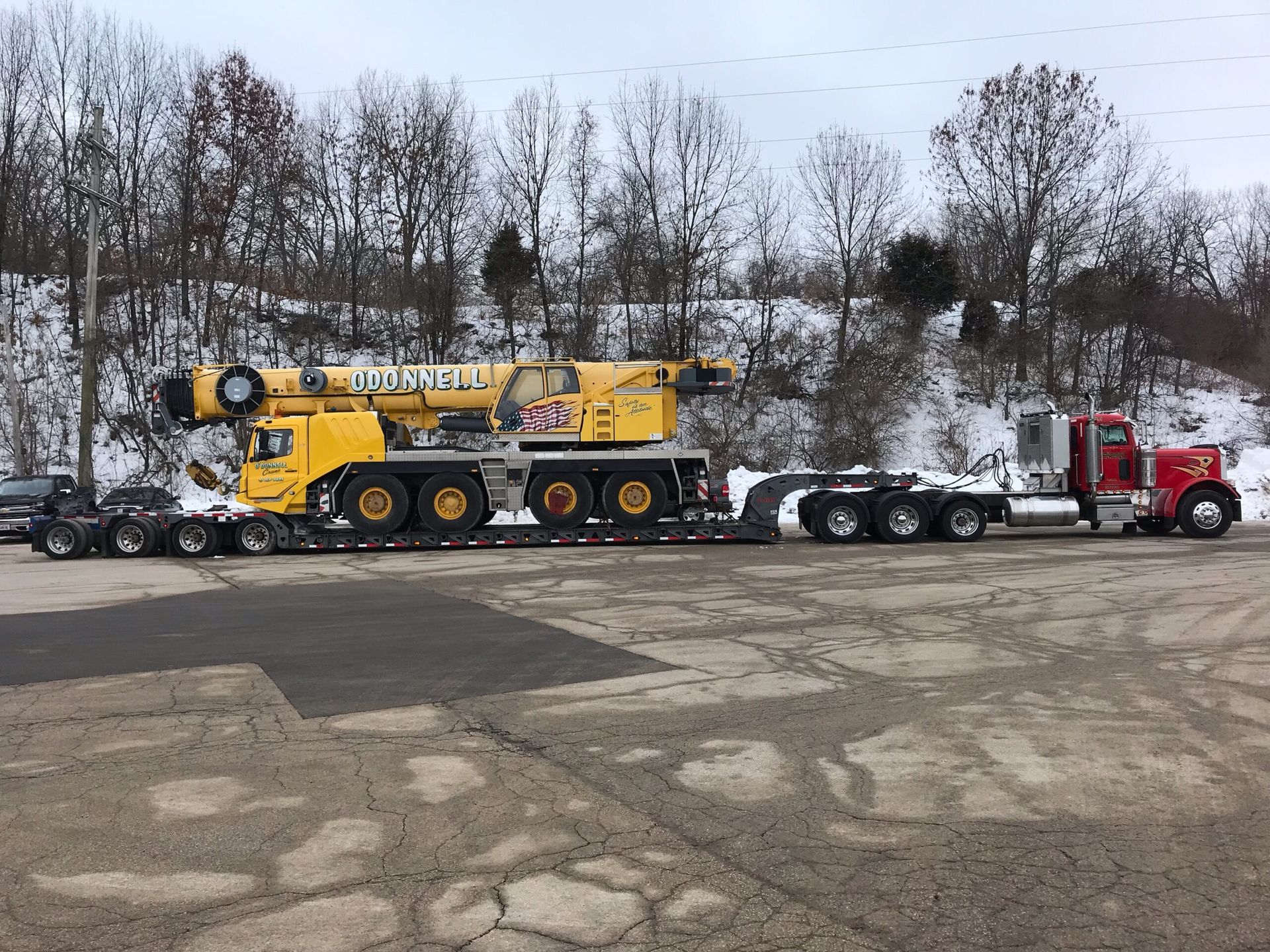 Yellow heavy crane on a red semi-truck in a snowy lot with bare trees in the background