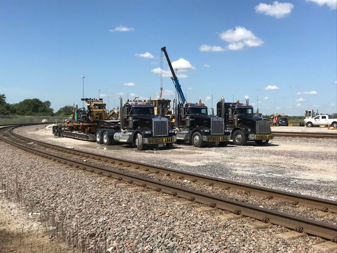 Row of black construction trucks on a gravel lot beside railroad tracks under a blue sky