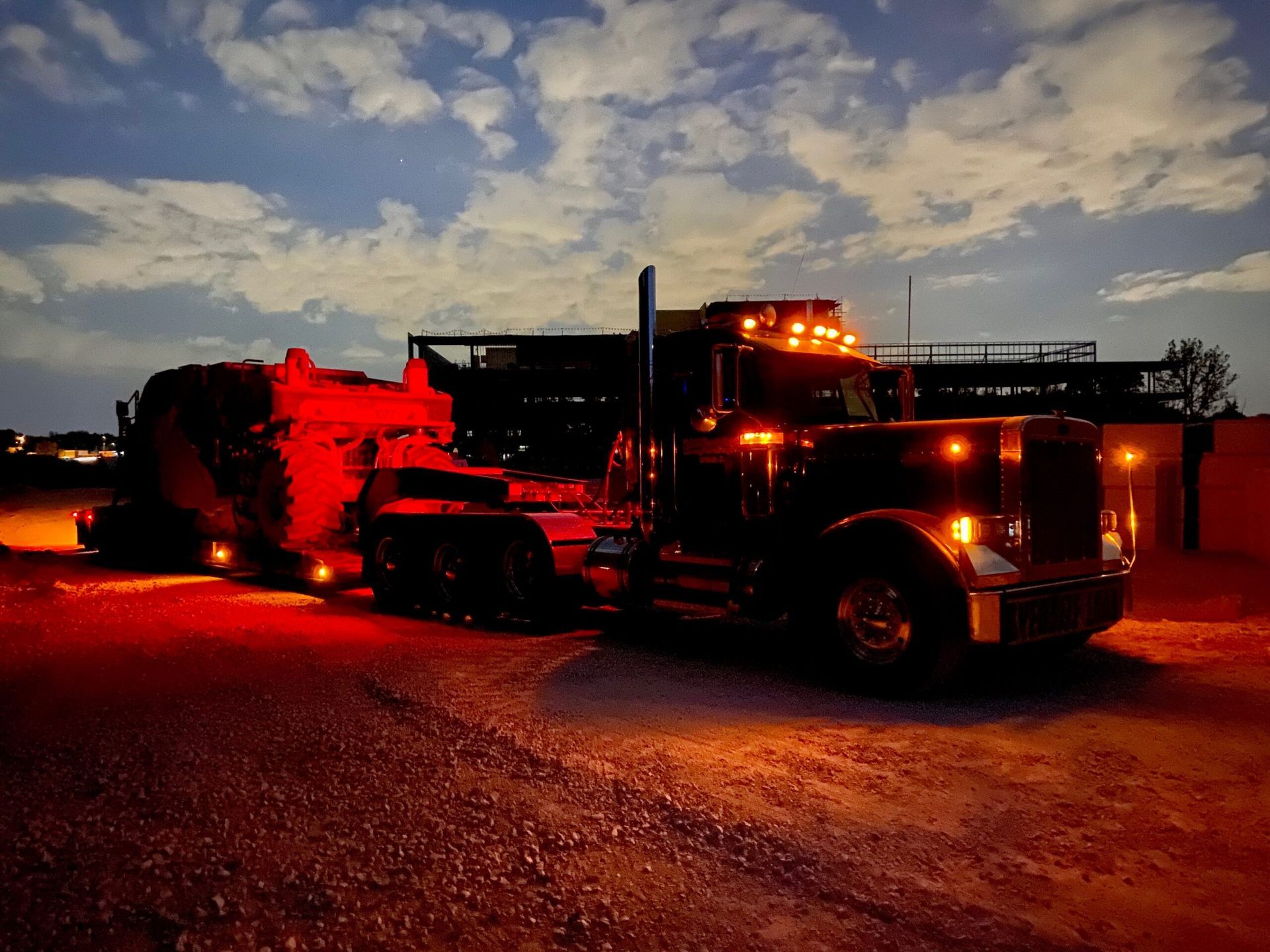 Red-lit semi truck parked at dusk on a gravel lot under a cloudy sky