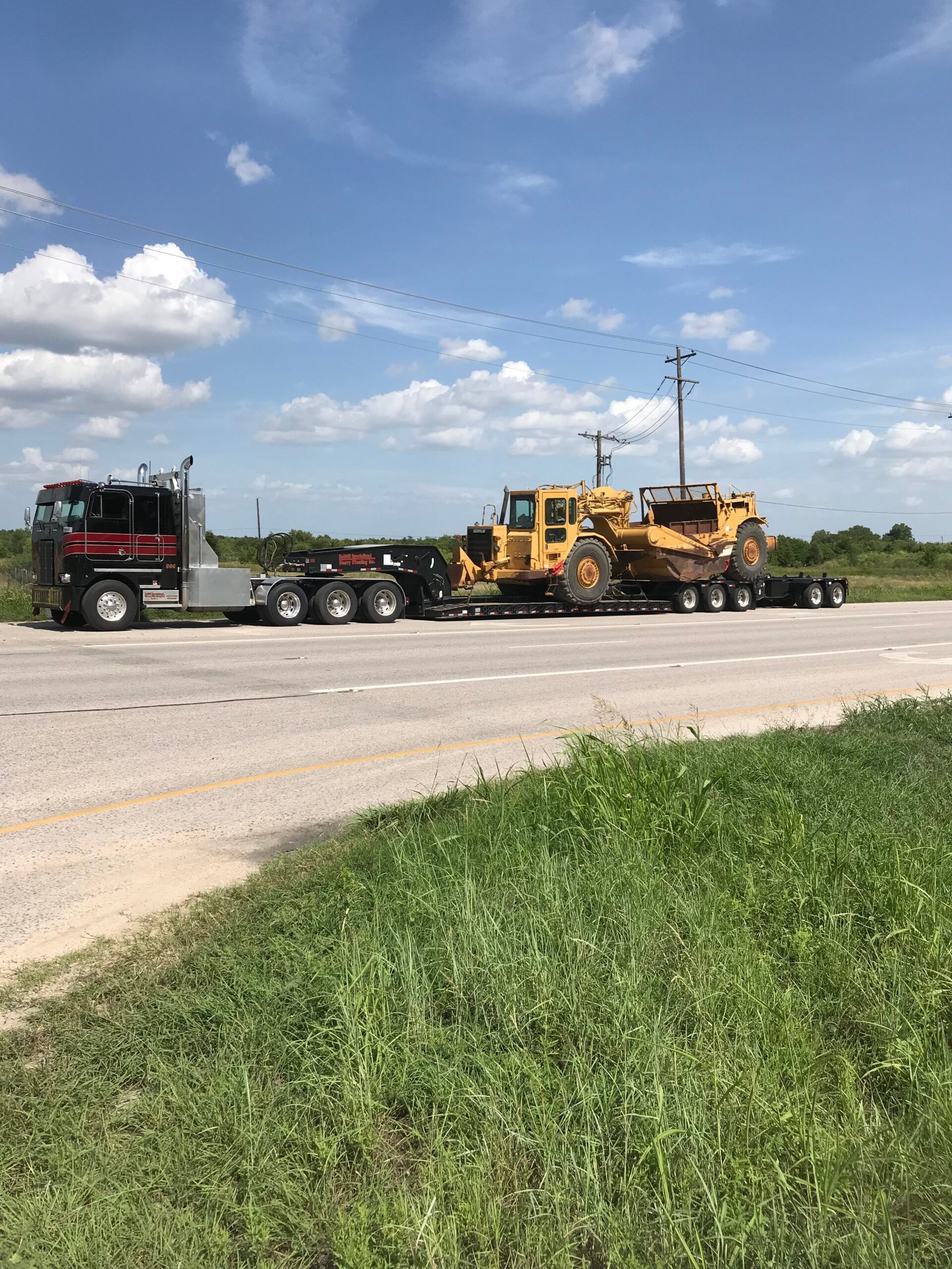 Heavy road construction machine towed by a black semi truck on a rural roadside under a blue sky