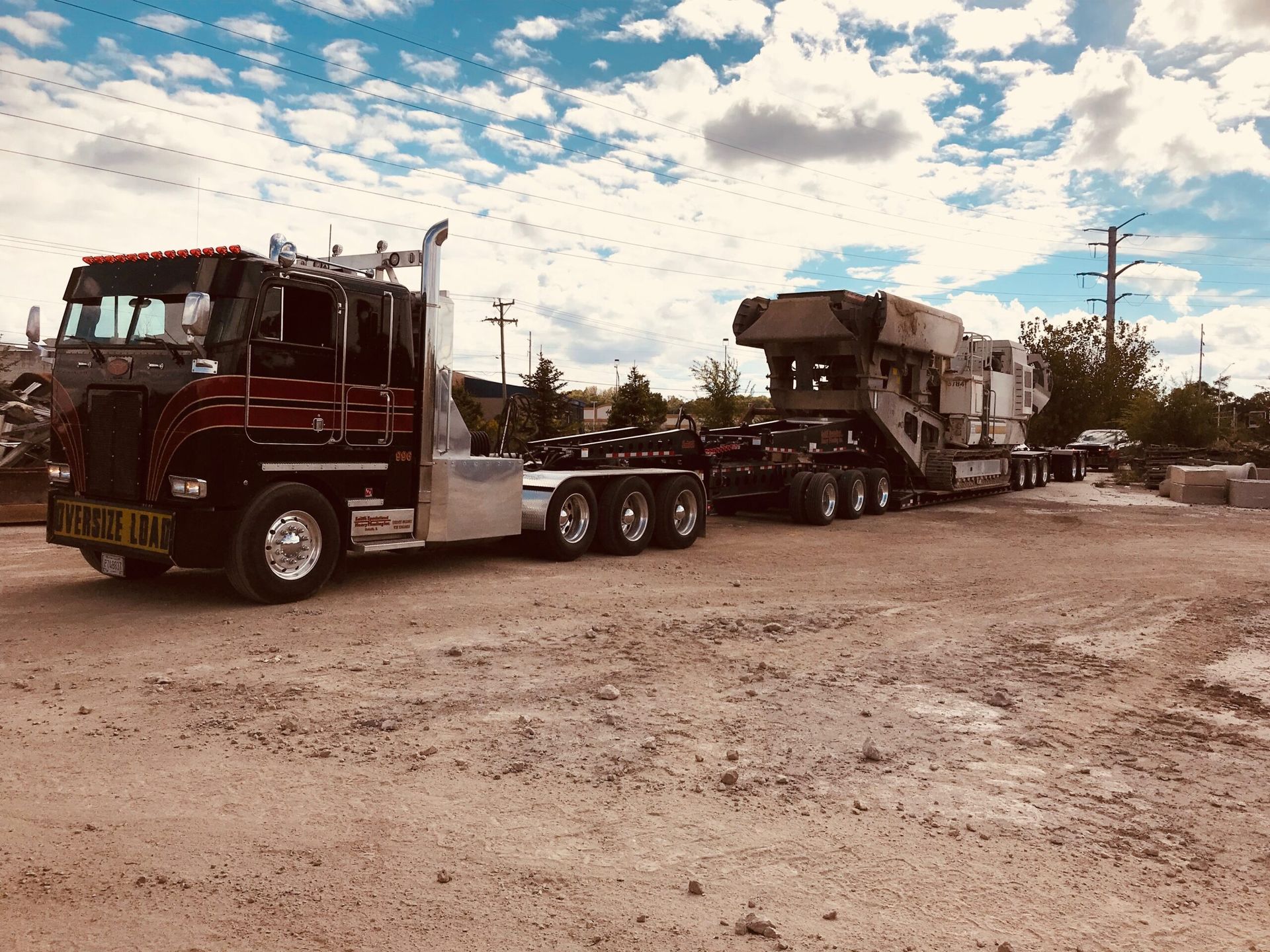 Semi-truck hauling a large piece of heavy equipment on a dirt road under a cloudy sky