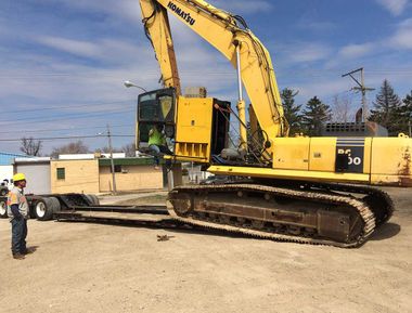 Yellow excavator on tracks parked on a dirt lot, with a worker standing nearby under a blue sky.