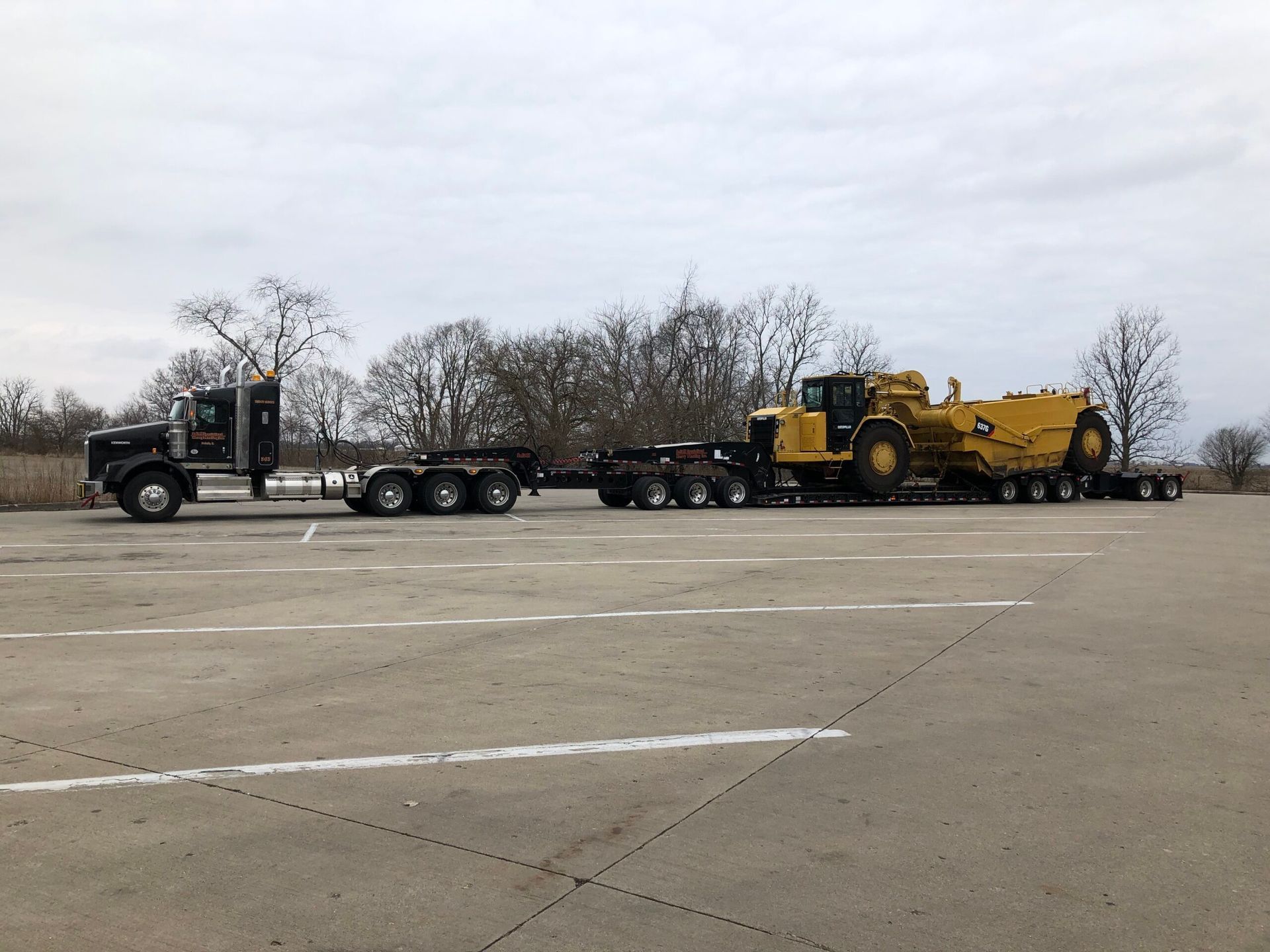 Black semi-truck hauling a yellow construction machine on a multi-axle trailer in a parking lot.