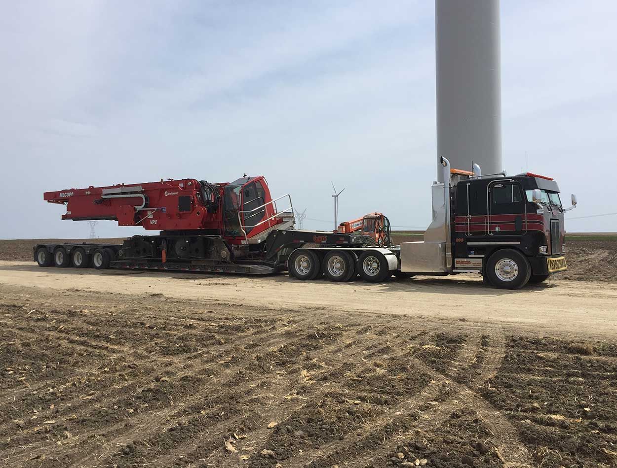 Red crane on a flatbed truck parked beside a wind turbine in a field