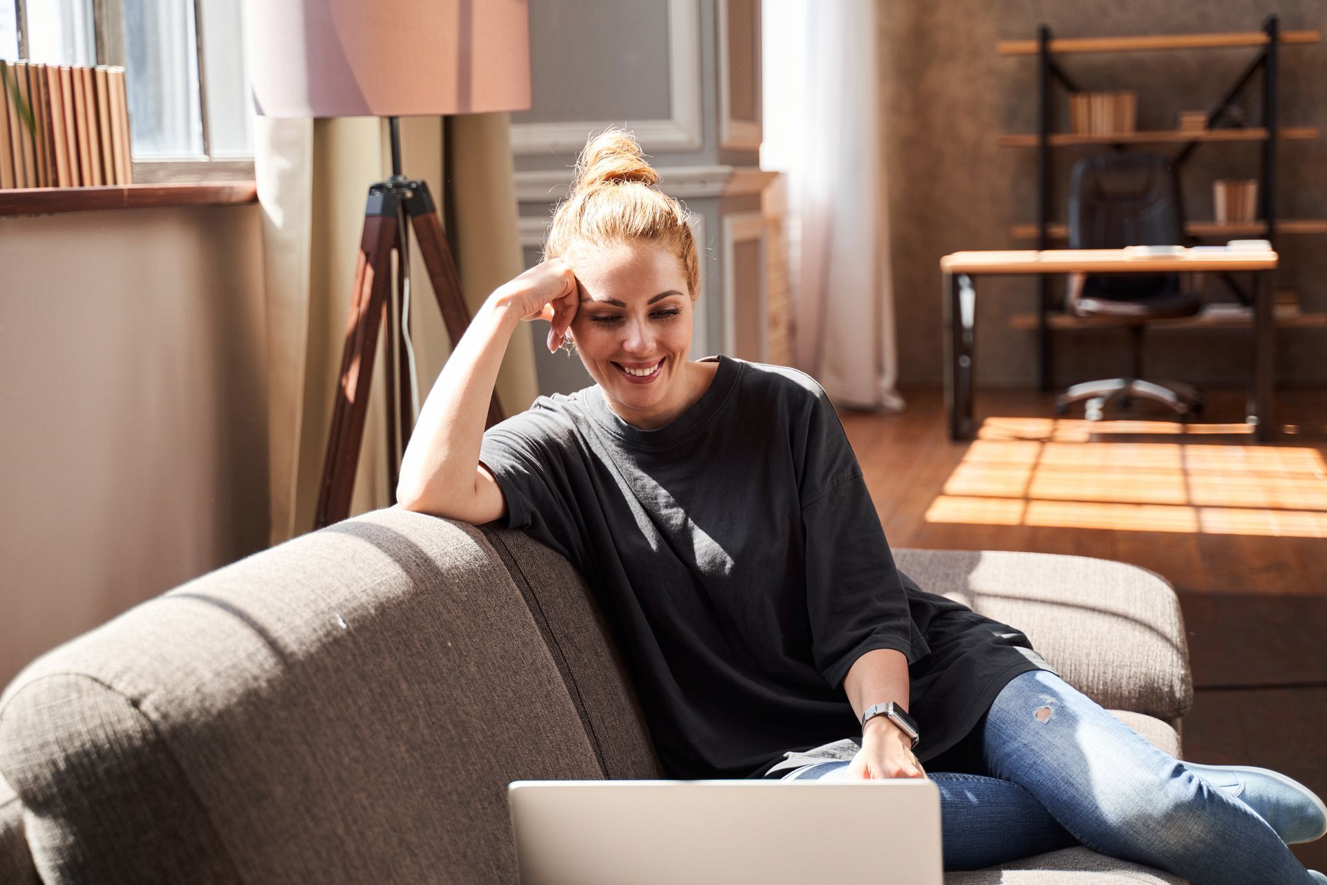 Woman smiles while using laptop on a couch in a sunlit room.