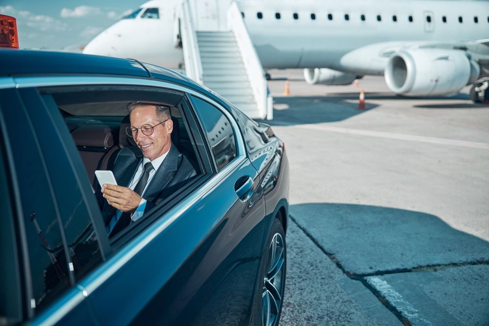 Man in suit smiles, looking at phone in car by airplane. Airport tarmac setting.