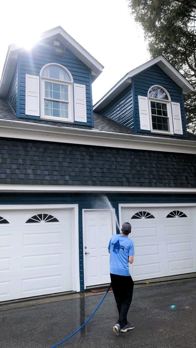 Person standing on a wet driveway spraying a blue house with a pressure washer.