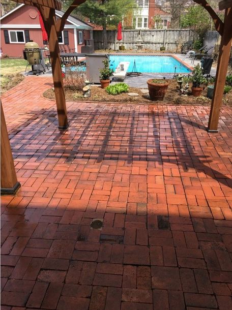 Red brick patio with a wooden pergola, leading to a blue swimming pool in a backyard.
