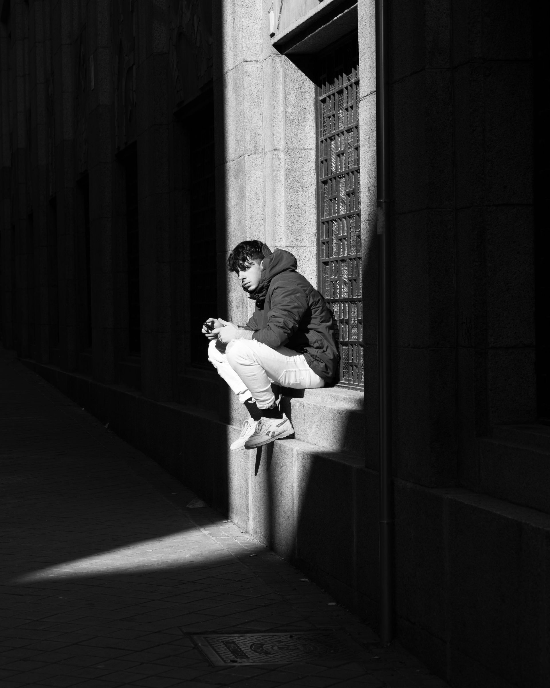 Person sitting on a sunlit ledge in a dark alley, reading a book in black and white.