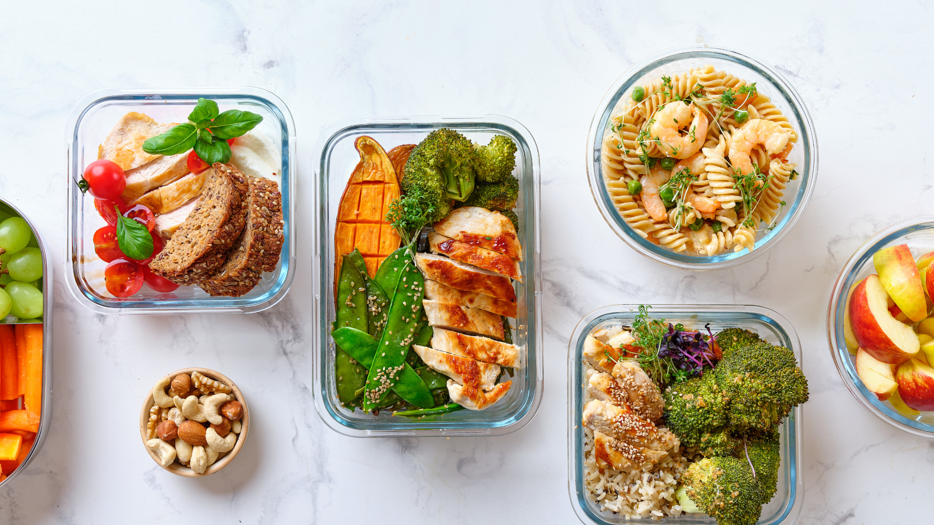 Meal-prepped food in glass containers: chicken, pasta with shrimp, vegetables, and snacks on a white surface.
