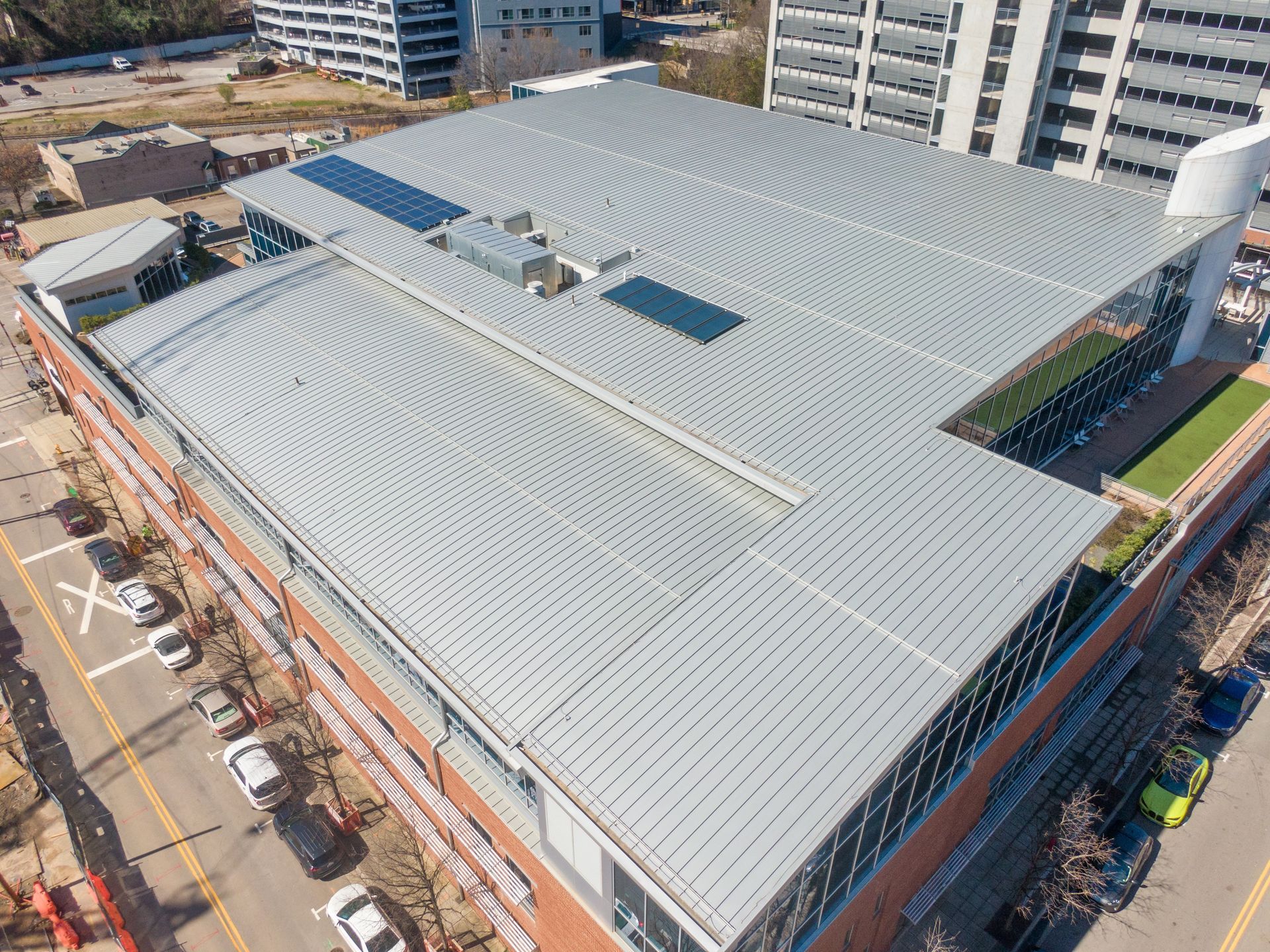 Aerial view of a modern building with a metallic, patterned roof, next to a street with parked cars.
