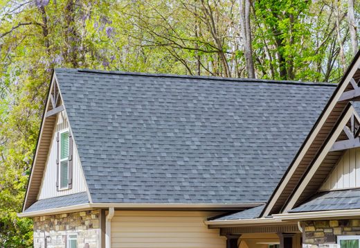 Gray shingled roof of a house with light tan siding, trimmed in brown, and a wooded background.