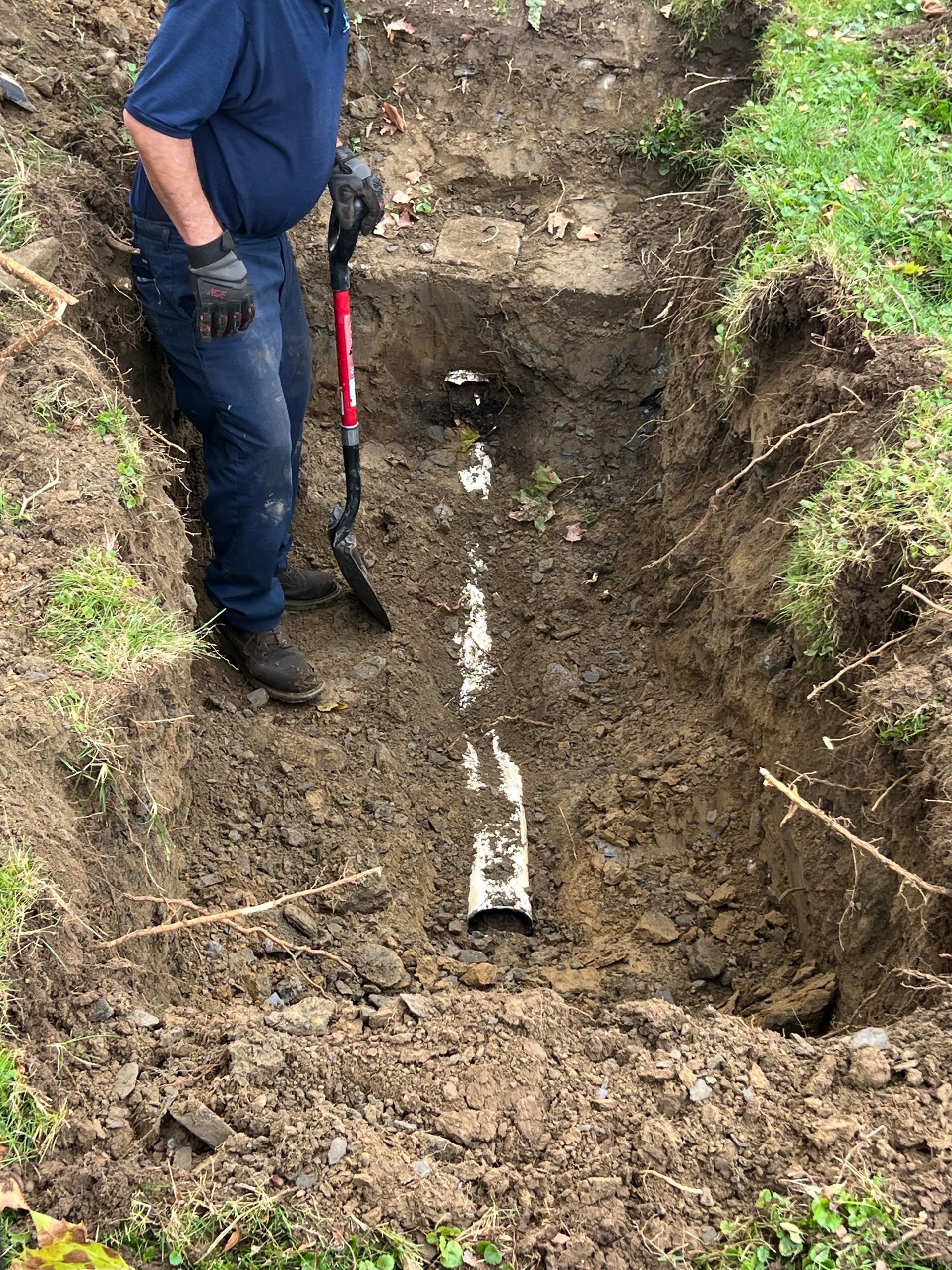 Person with red gloves using a rod to clear a drain in a grassy area.