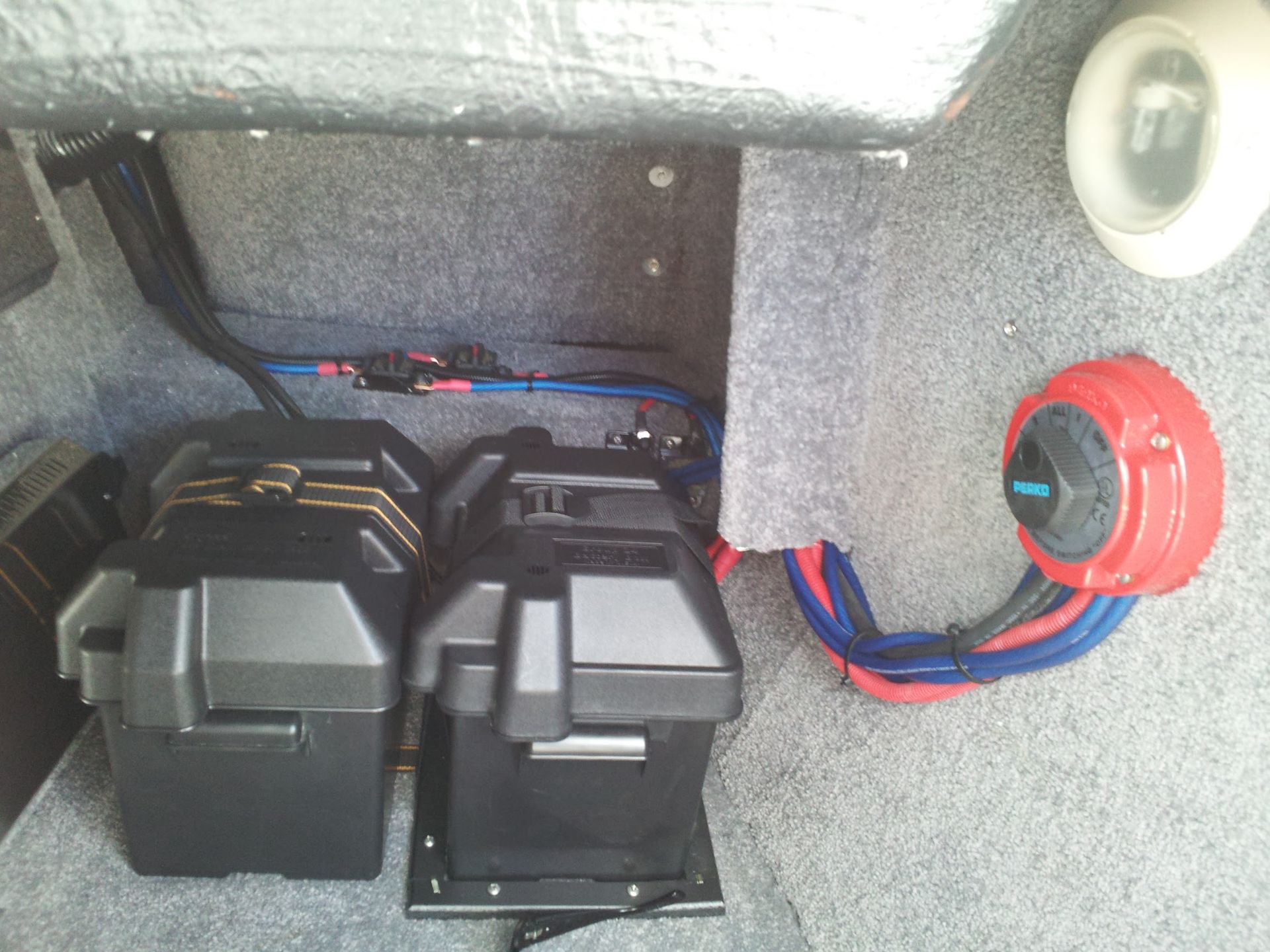 Car trunk interior with black battery boxes and wiring on gray carpet, plus a red circular connector.