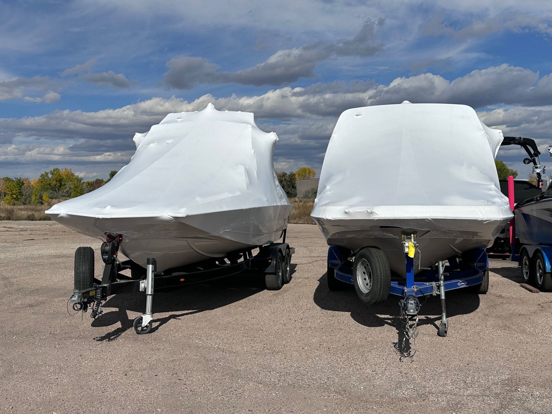 Two covered boats on trailers parked on gravel under a cloudy sky