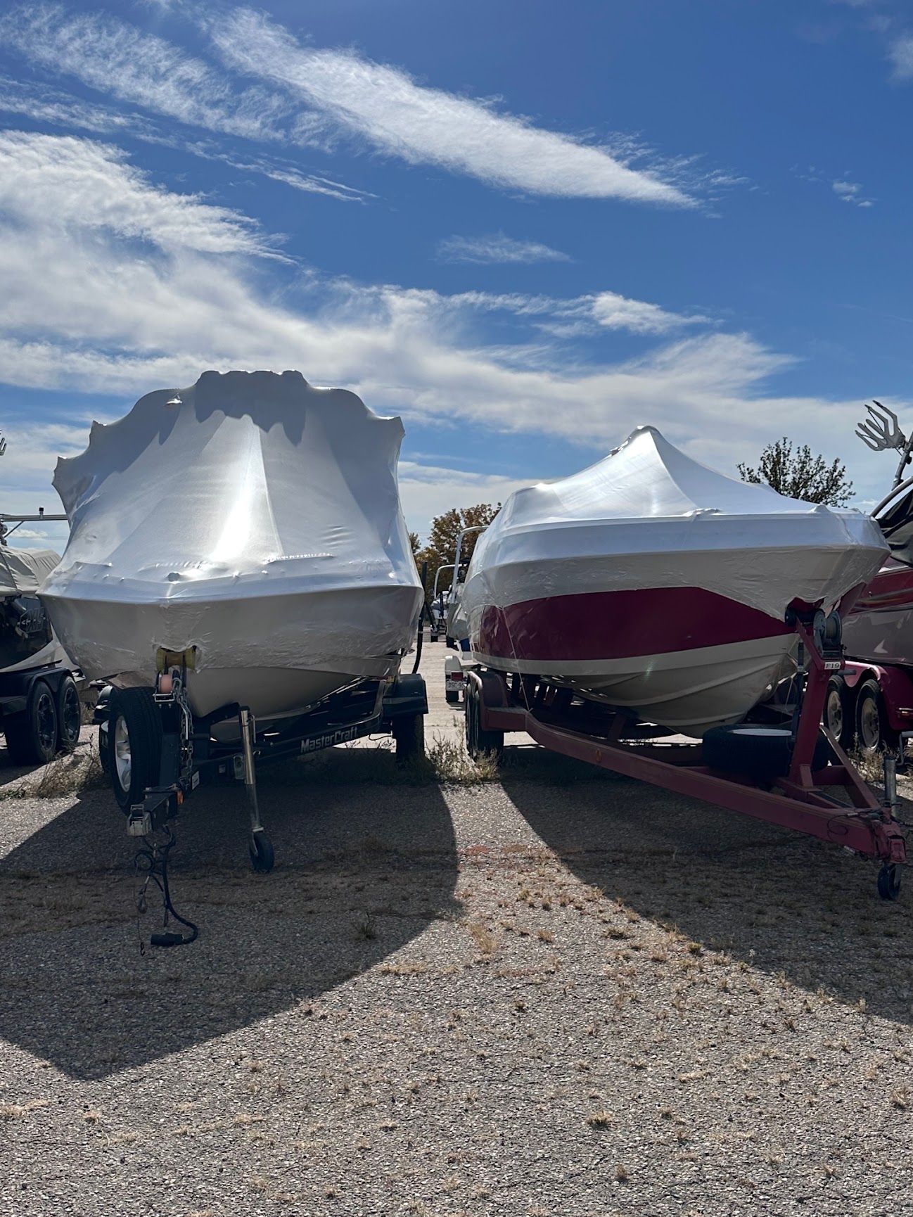 Two covered boats on trailers in a gravel lot under a blue sky