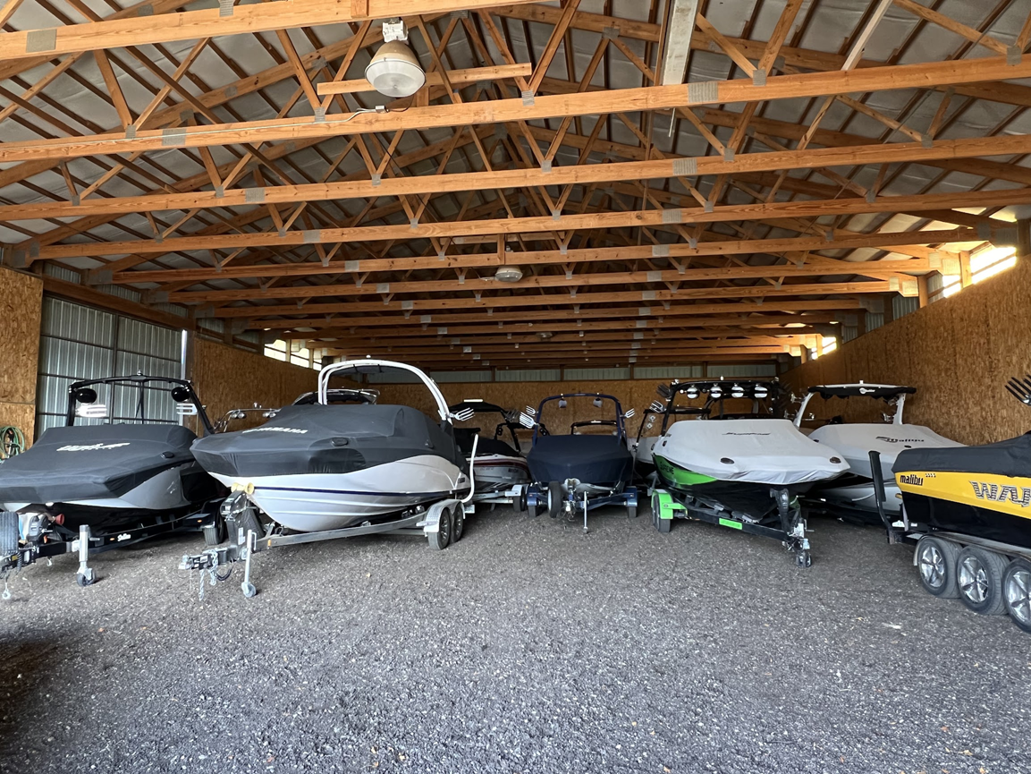 Several jet skis and a trailer stored in a gravel-floored garage with exposed wooden rafters.