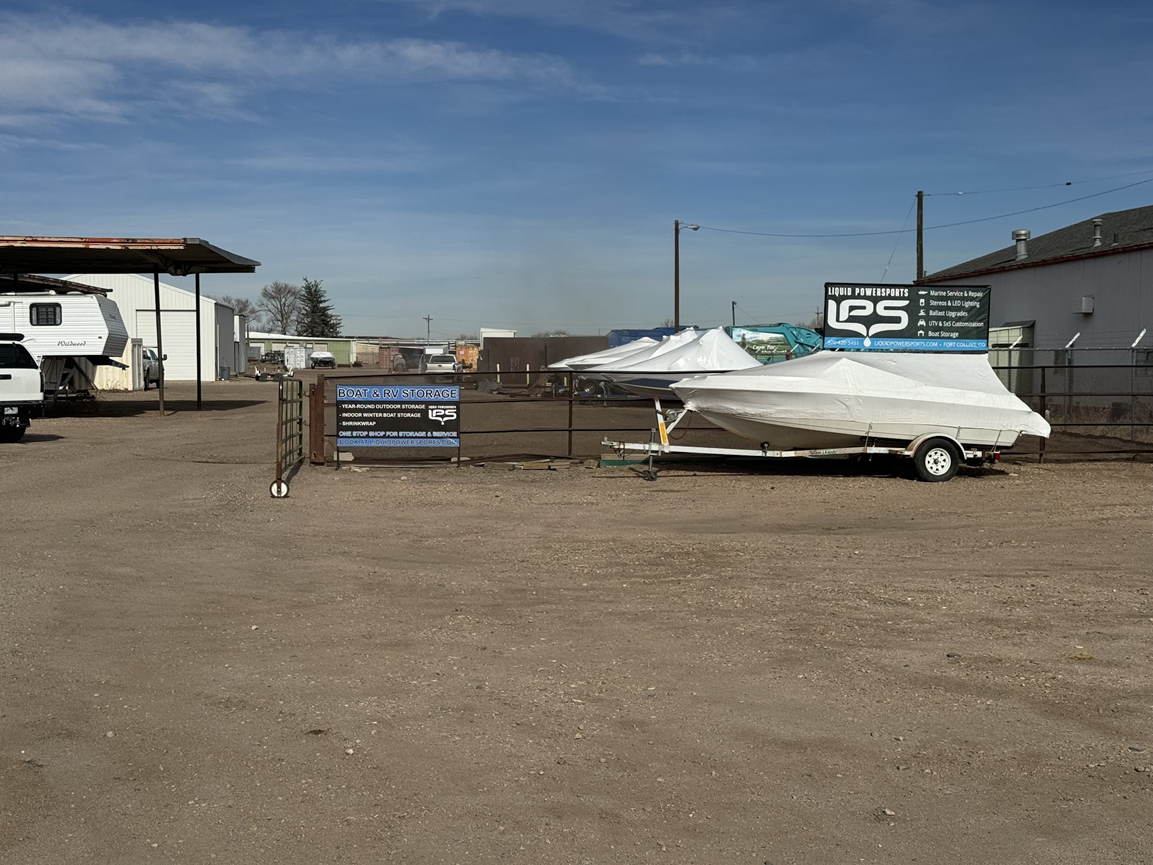 Outdoor lot with a covered white boat, trailers, and industrial buildings on a gravel surface.
