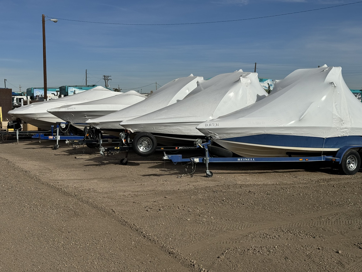 Rows of white covered boats on trailers parked in a gravel lot under a clear blue sky.