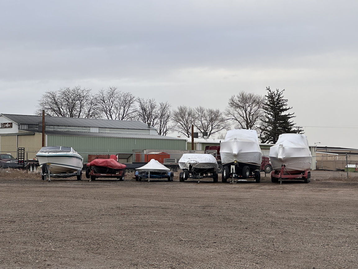Six boats stored on trailers in a gravel lot under an overcast sky
